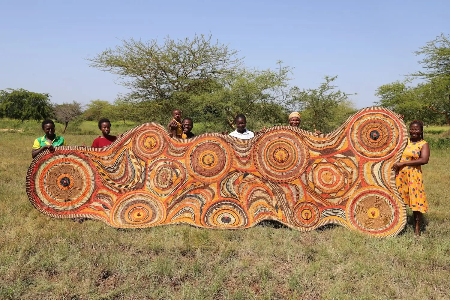 Ghanaian artisans stand in a meadow with a huge textile piece woven from PET bottles
